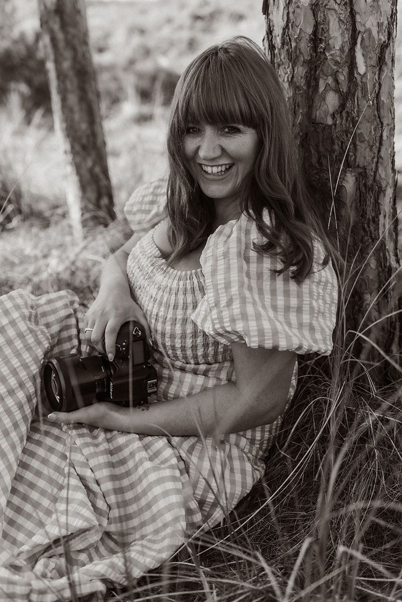 Black and white photo of female photographer in gingham dress holding camera while sitting against tree in Lancashire location