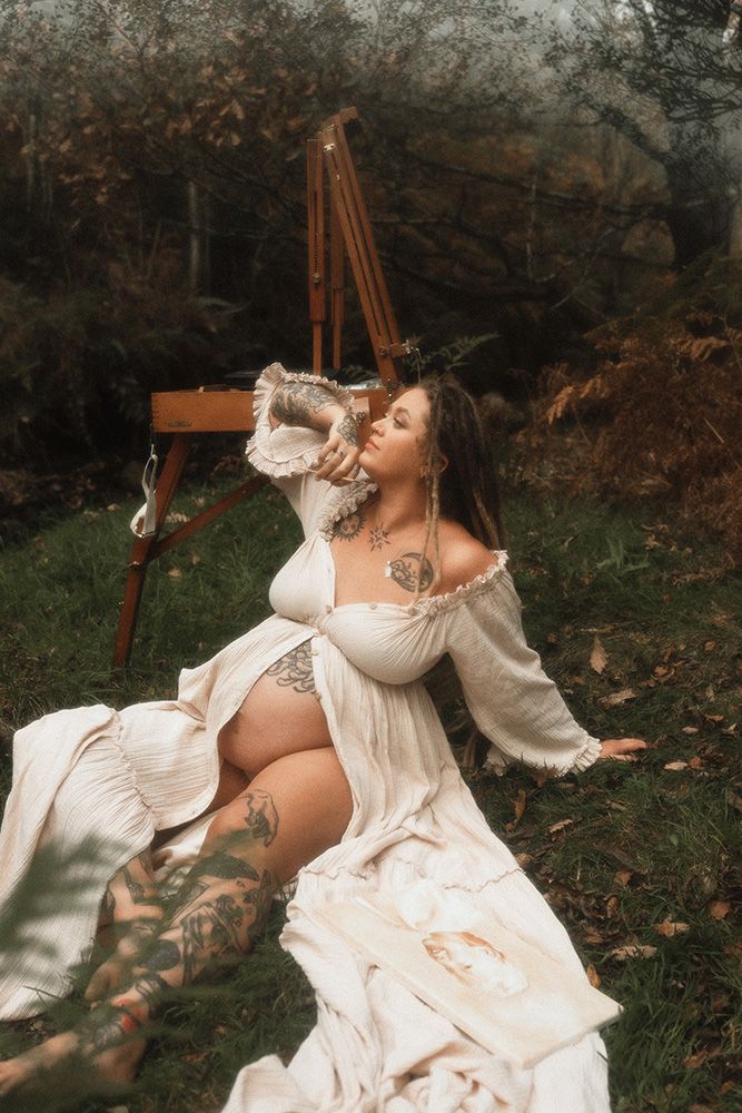 Manchester maternity photographer. Pregnant woman in white boho dress with tattoos sits outdoors near an easel surrounded by autumn foliage.