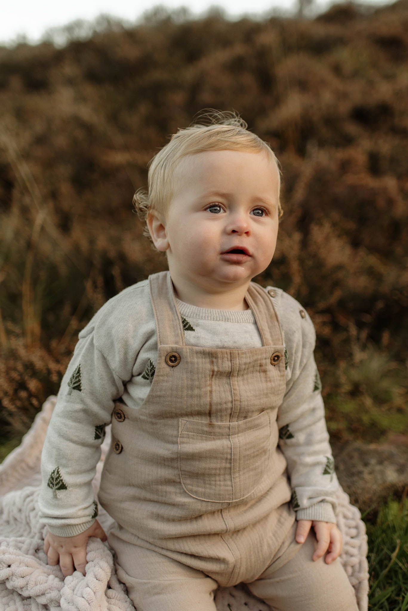 A young child wearing beige overalls and a patterned sweater sits outdoors against a natural autumn background during Chorley family photos
