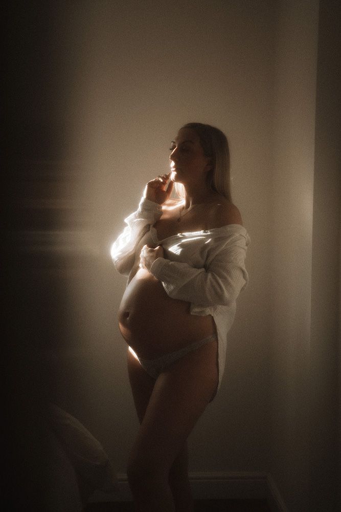 Manchester maternity photographer captures pregnant woman in white off-shoulder top and underwear cradling baby bump in soft moody light.