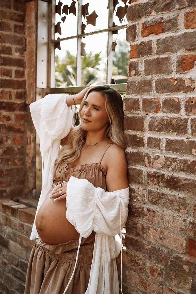 Pregnant woman in brown dress and white cardigan posing against rustic brick wall during maternity photoshoot.