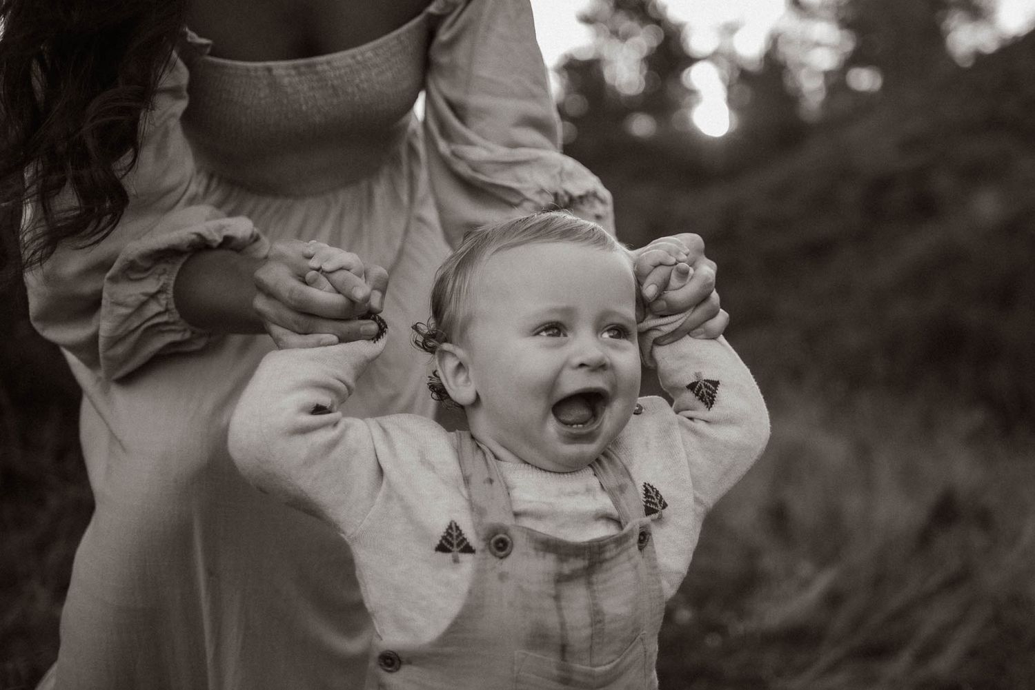 A joyful baby laughs while being supported by someone holding their arms in a black and white photograph.