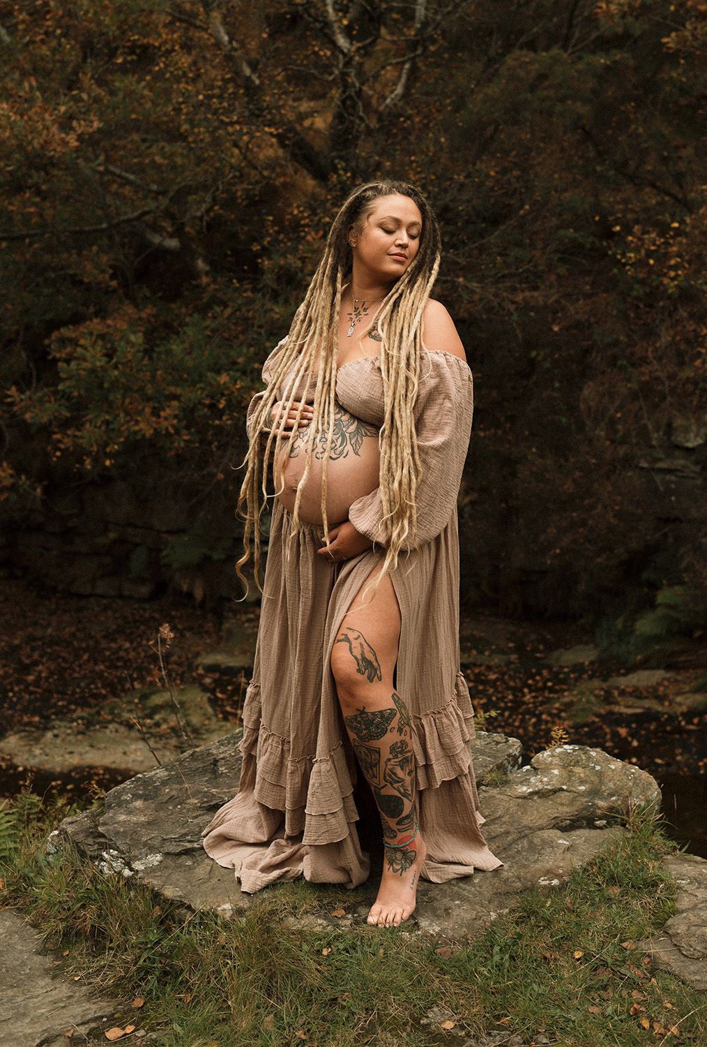 A maternity photoshoot in Lancashire, in nature shows a figure in flowing beige dress seated on rocks against an autumn forest backdrop.