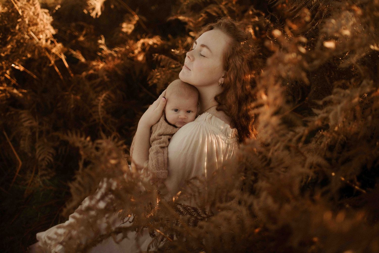 A tender moment captured in sepia tones shows a parent and baby embraced among golden autumn foliage.