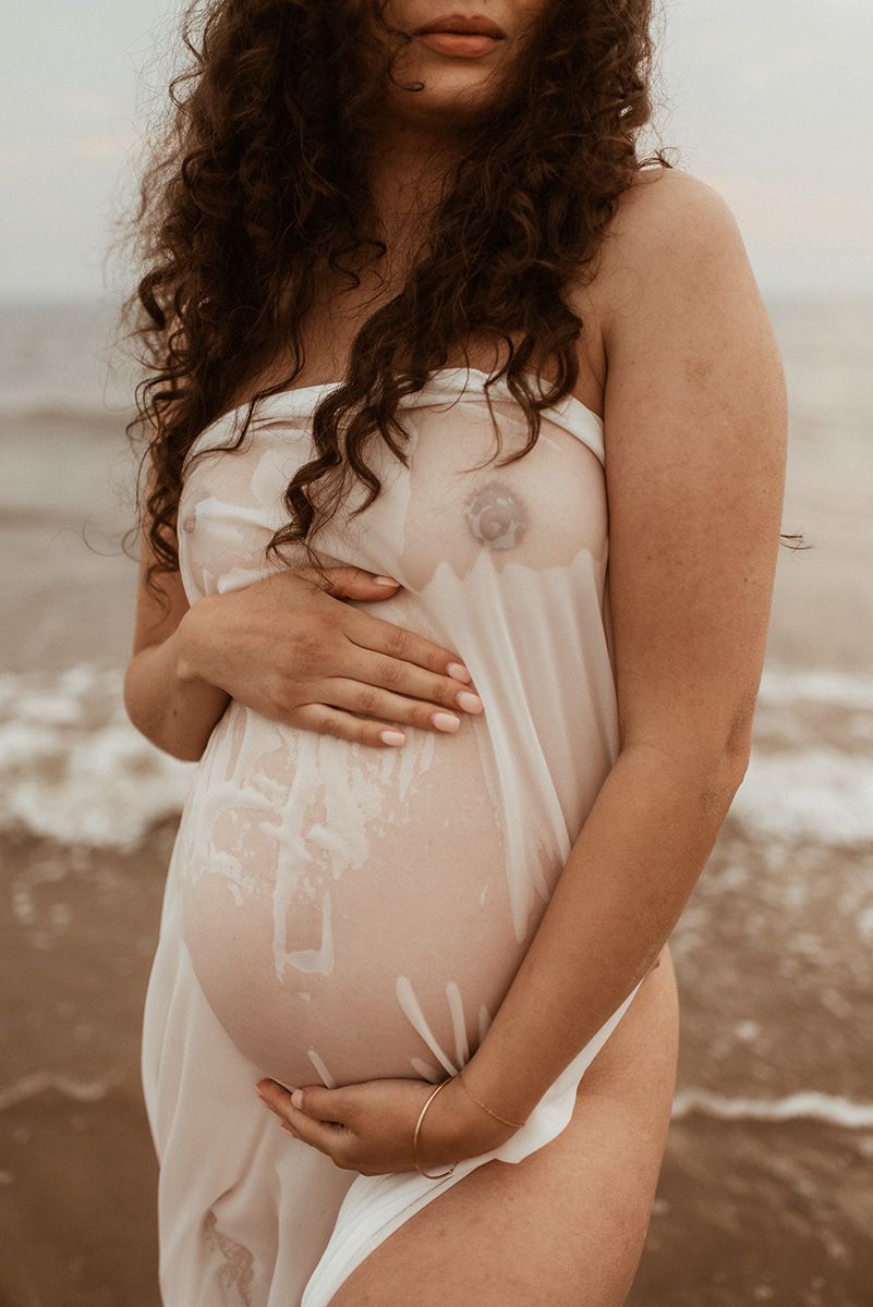A pregnant woman in a white dress stands on a beach while lovingly holding her baby bump during a maternity photoshoot in Lancashire