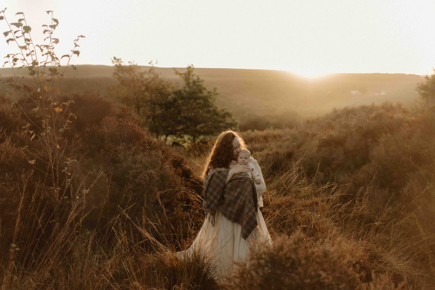 A person wrapped in a blanket stands on a hillside at golden hour overlooking a misty rural landscape.