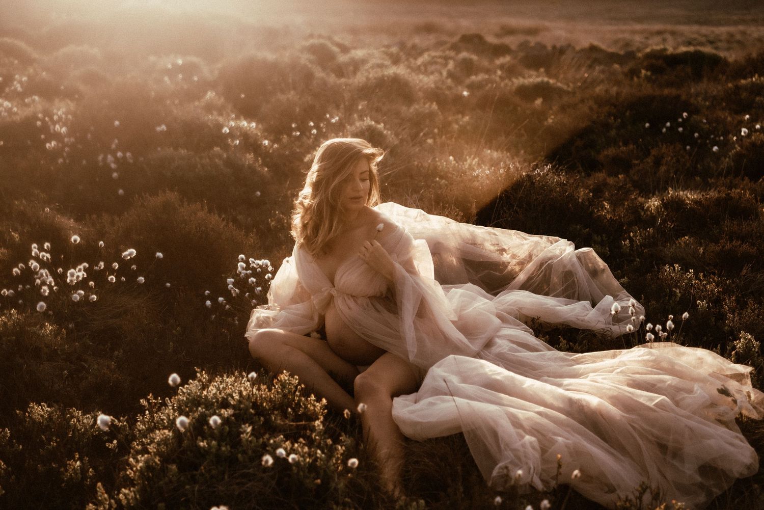 Ethereal figure in flowing white dress lies amongst wildflowers in dreamy golden sunlit meadow at dusk.