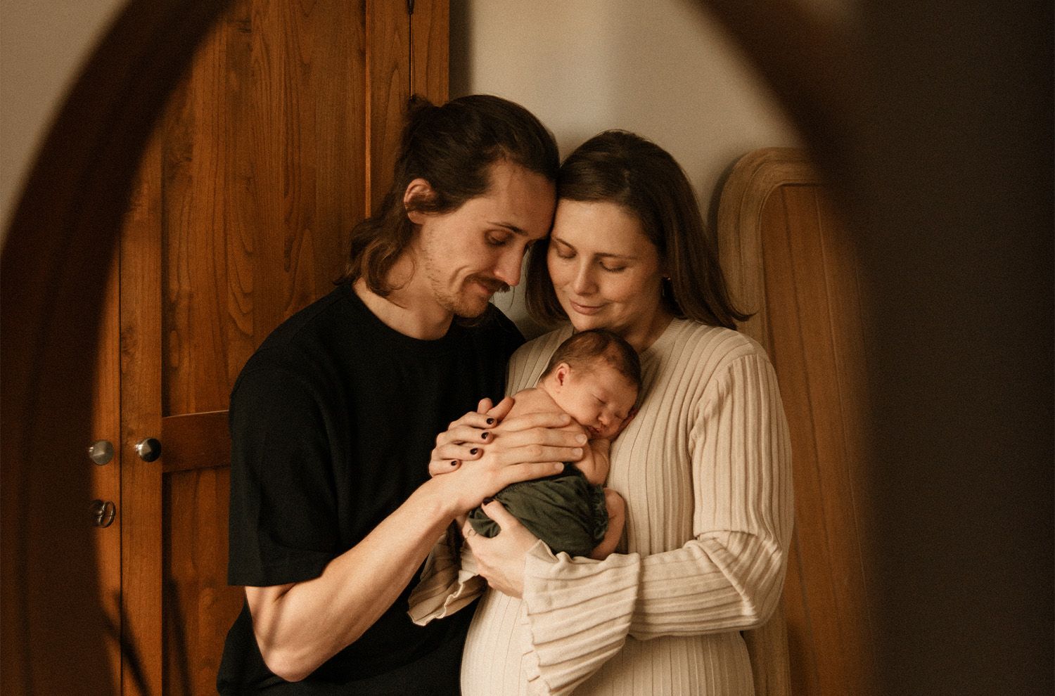 A couple embraces in a warmly lit rustic wooden interior setting, with their newborn. on a photoshoot at home in Manchester.