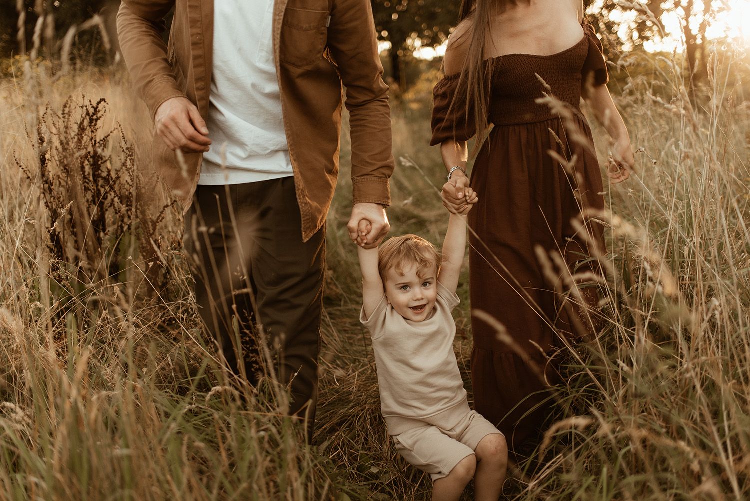 A joyful young child holding parents hands while playing in a tall grass field at golden hour during family photoshoot in Chorley