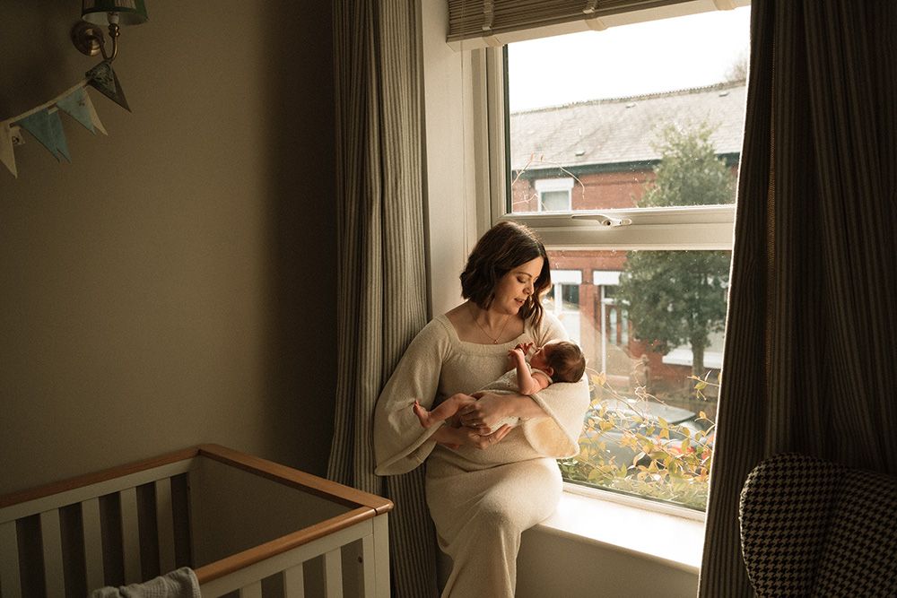 Newborn photographer in Manchester captures  photos showing a person by a window holding and feeding a baby in natural light.