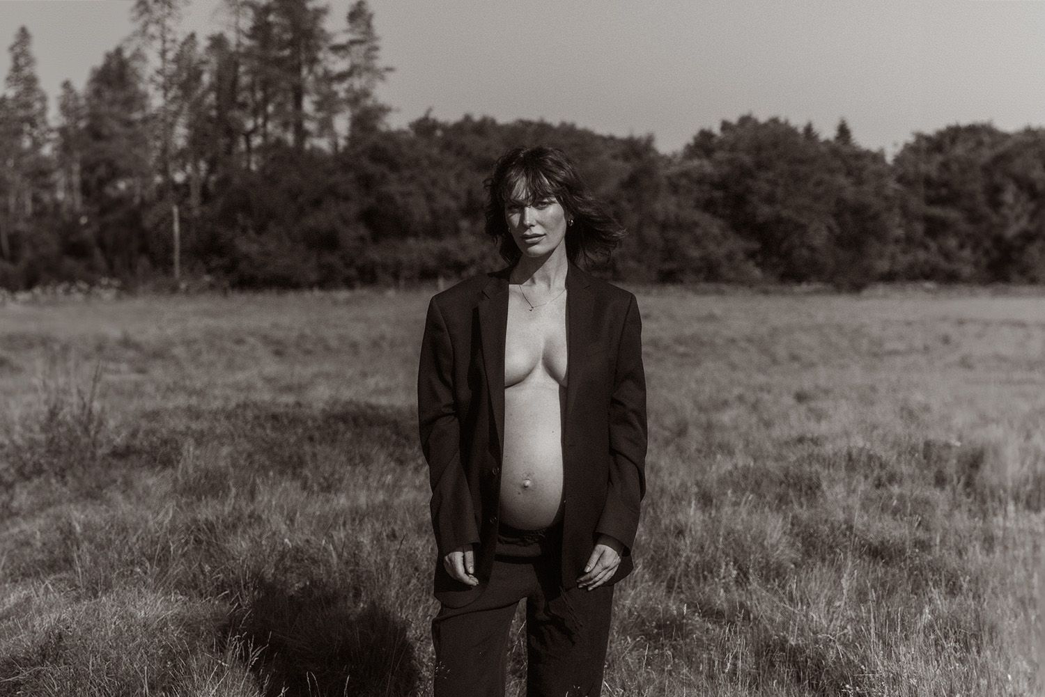 A black and white portrait shows a pregnant woman in a dark jacket standing in a grassy field with trees in the background.