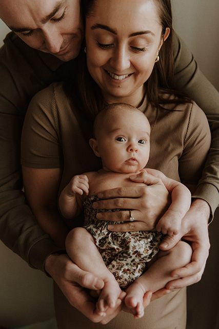 A tender family portrait shows parents in brown clothing holding their baby in a floral-patterned outfit during newborn photoshoot Chorley