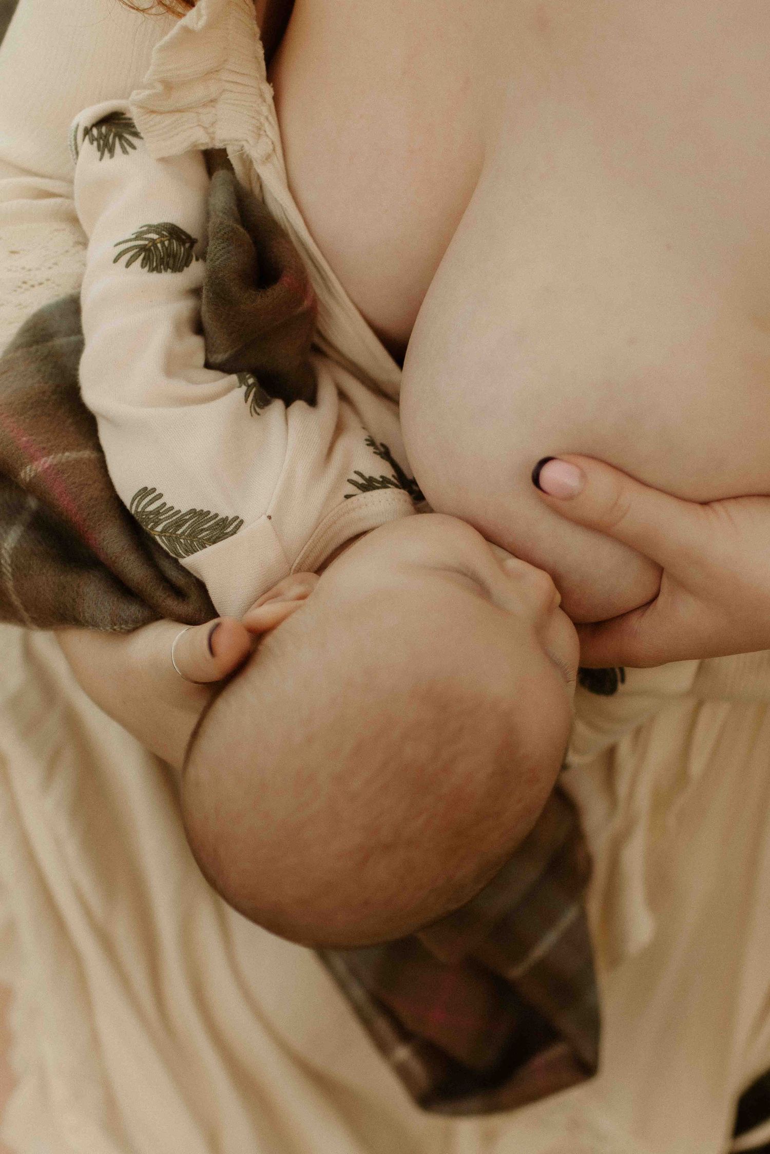 Intimate breastfeeding moment with baby wearing feather-printed outfit while laying on soft beige bedding.