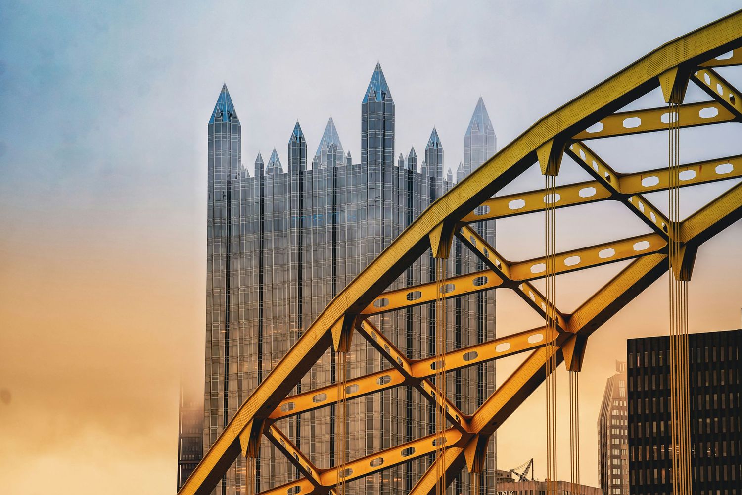 A yellow steel bridge arch frames Pittsburgh's PPG Place skyscrapers against a colorful sunset sky.