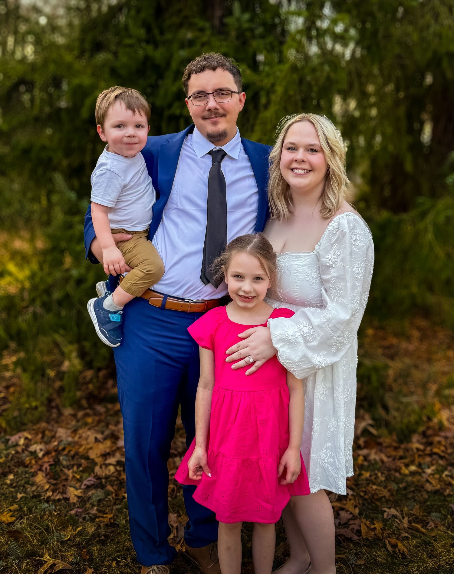 Family portrait outdoors showing parents with two children in casual formal attire against a natural background.