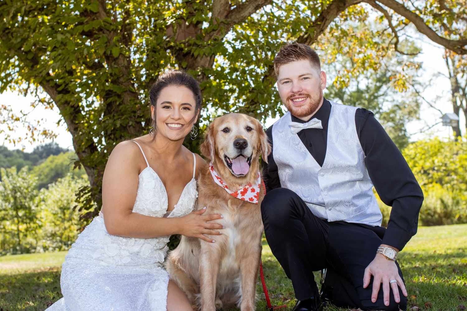 A couple in wedding attire poses outdoors with their golden retriever wearing a floral collar on their special day.