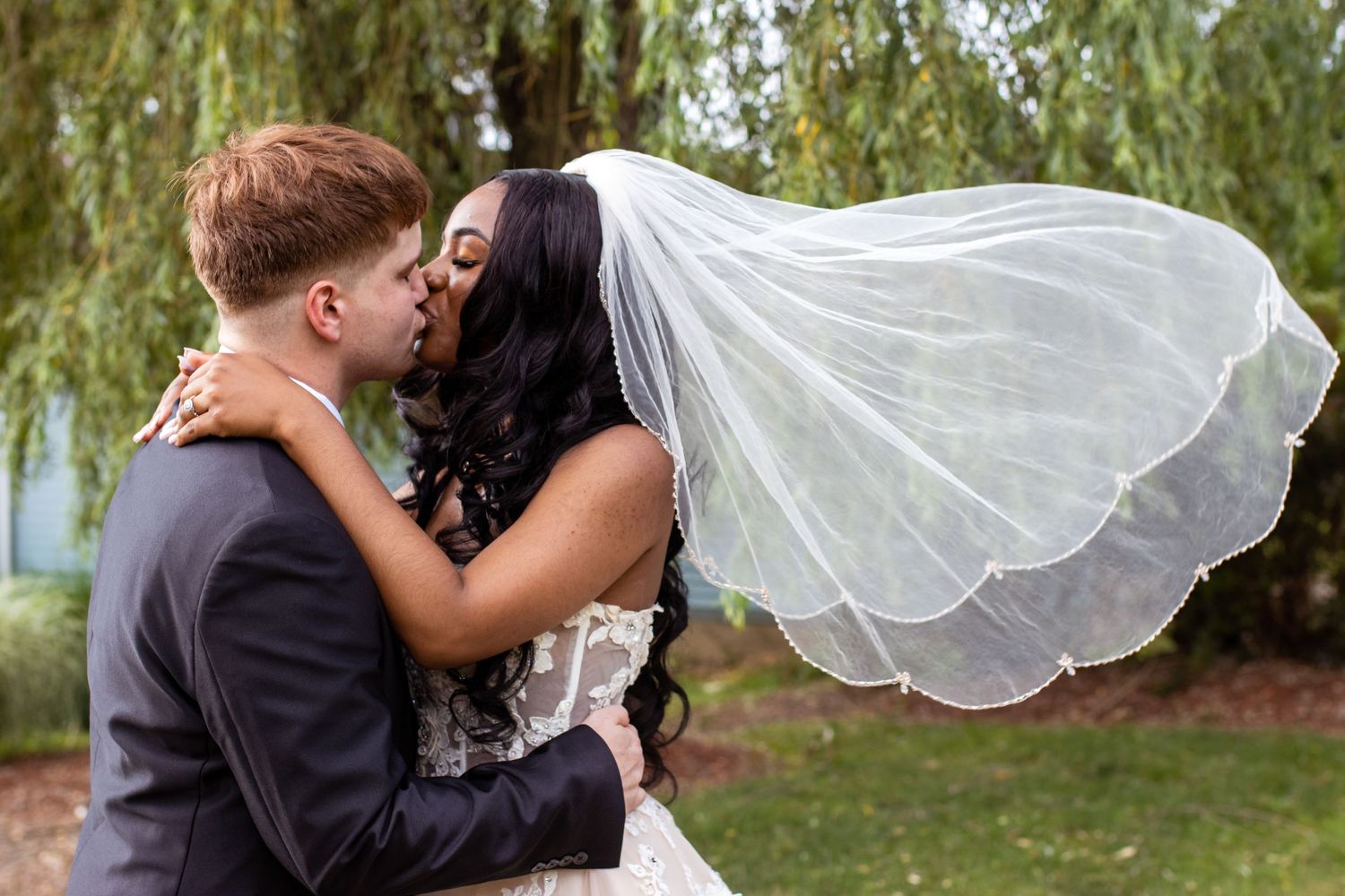 A romantic couple shares an intimate kiss outdoors with a flowing white bridal veil catching the breeze by a willow tree.