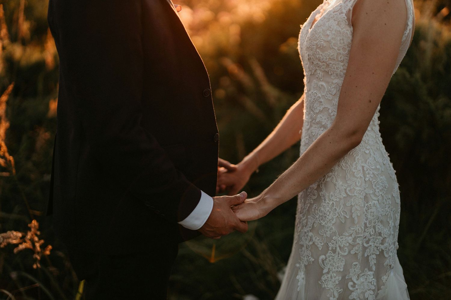 A couple holding hands at sunset, with bride wearing lace dress and groom in dark suit.