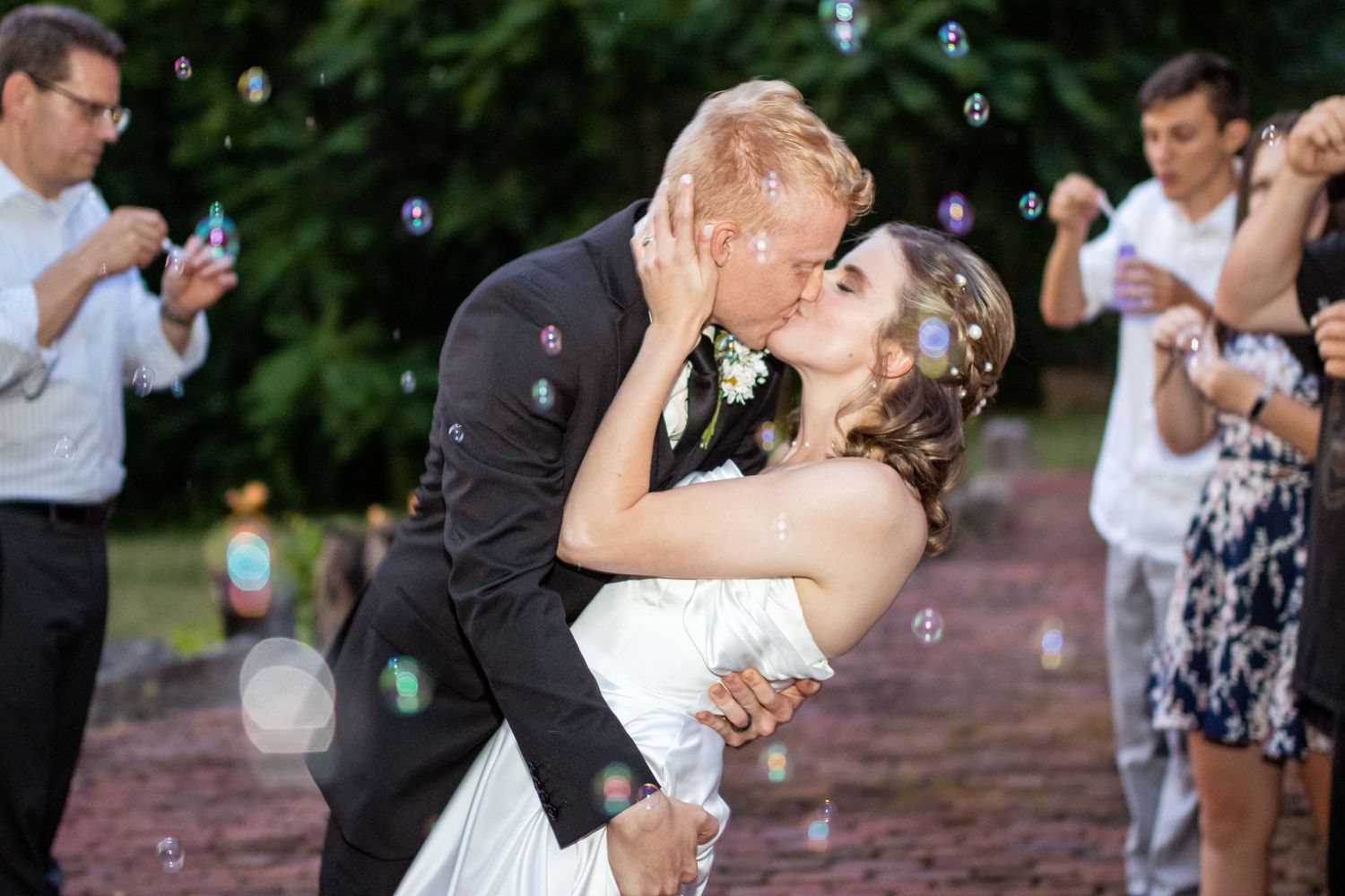 Newlyweds share an intimate kiss during bubble send-off while wedding guests celebrate in background at outdoor ceremony.