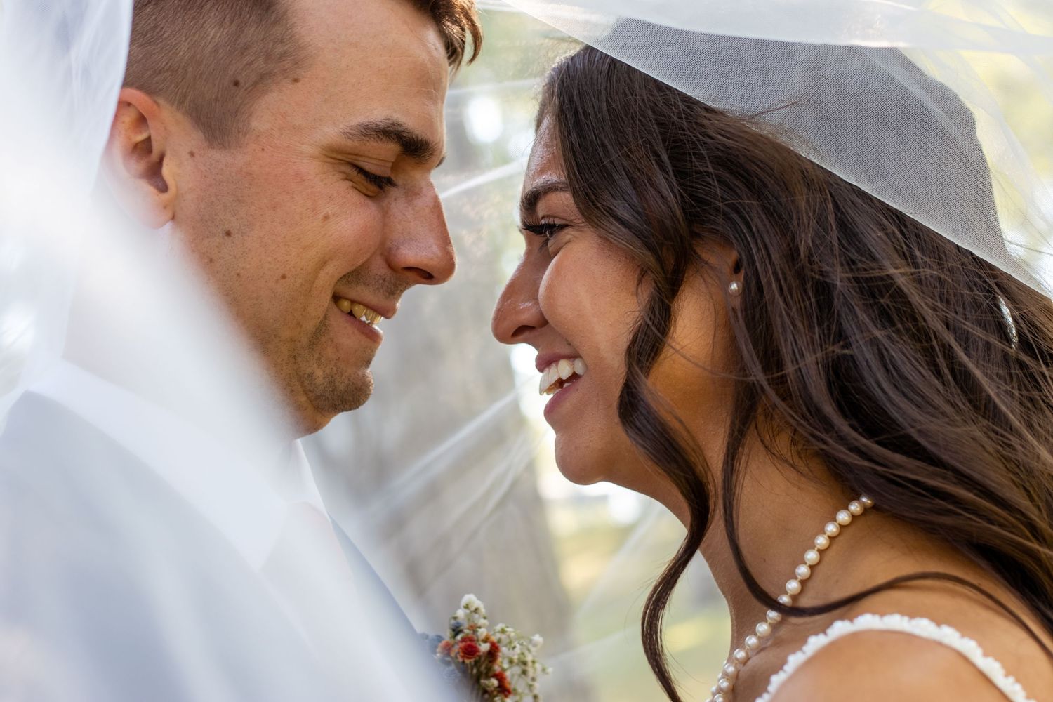 A close up of two people sharing an intimate moment under a wedding veil with white attire and pearls.
