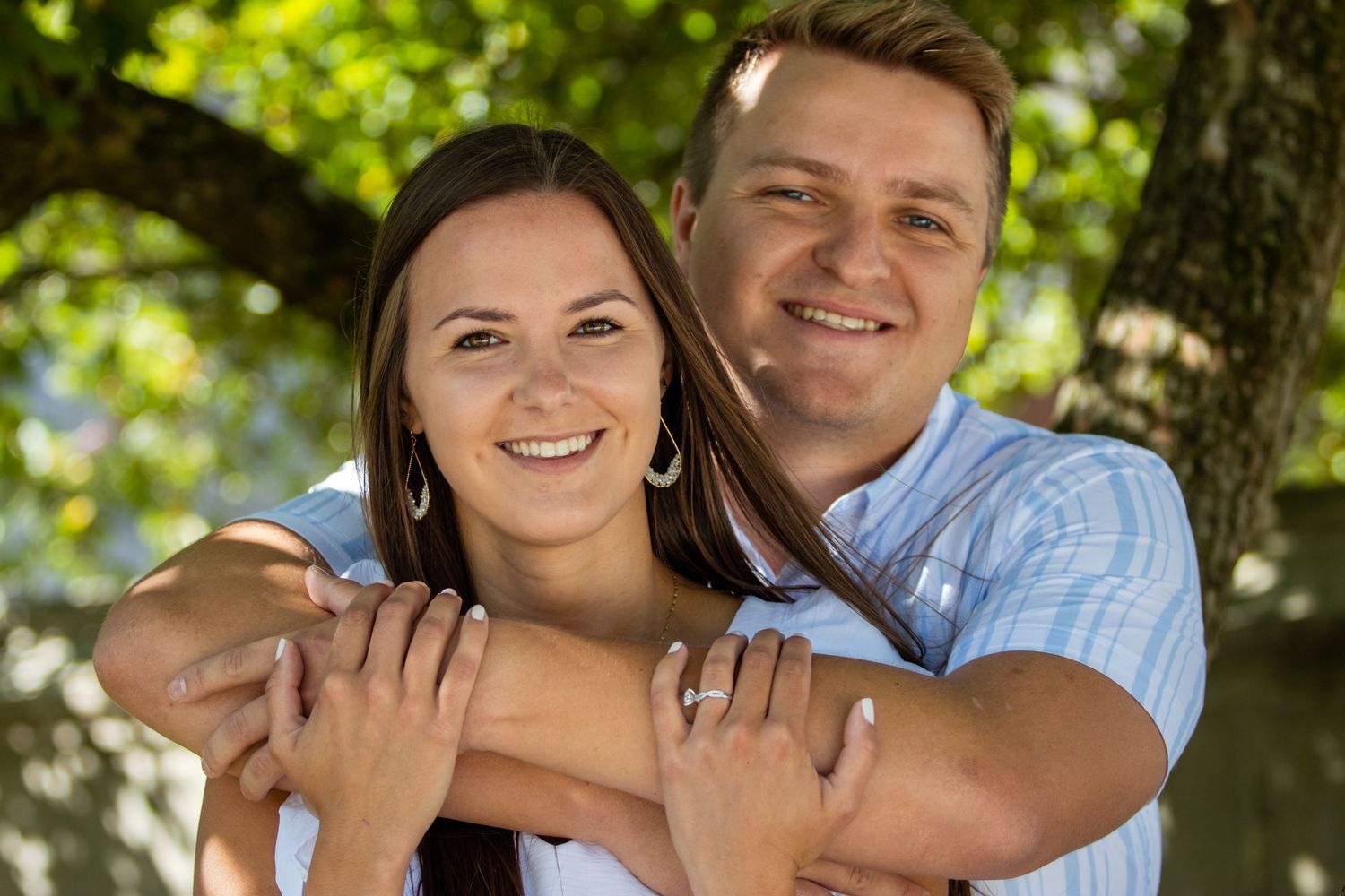 A couple hugs outdoors in front of a tree with green foliage in the sunlight.