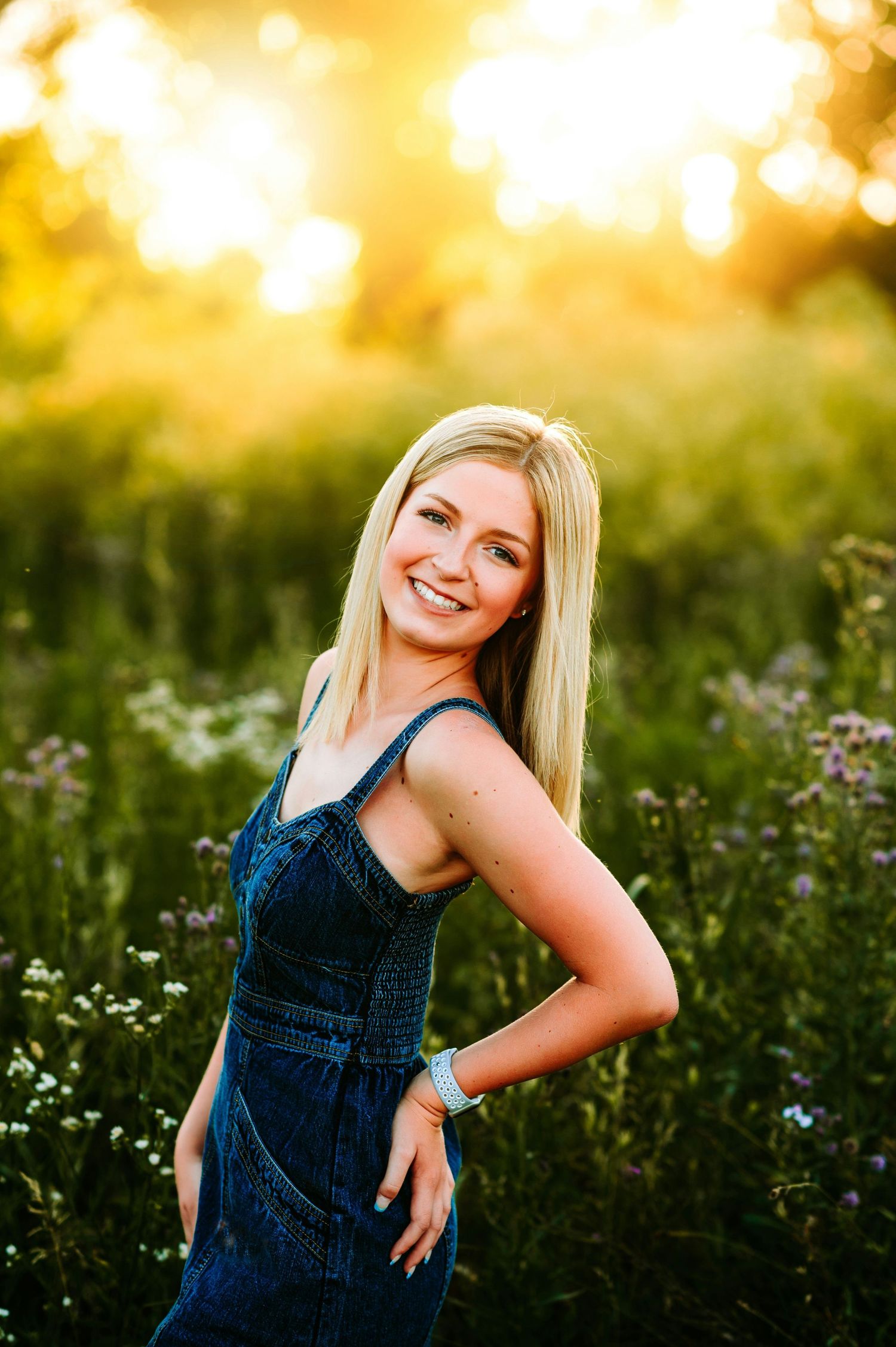 A figure in a blue denim dress poses in a wildflower meadow at golden hour.