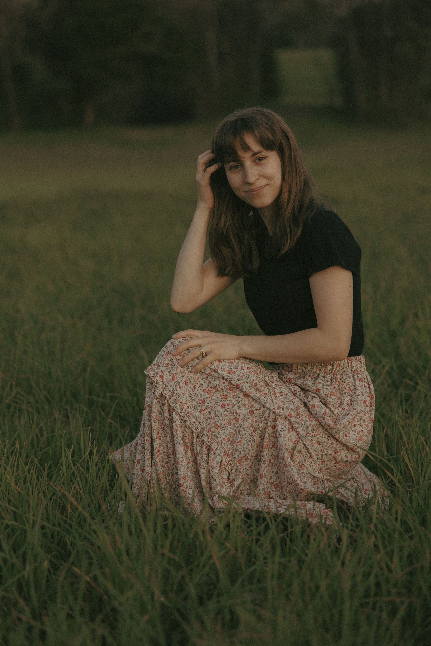Young woman in floral maxi skirt sits gracefully in lush green grass field.