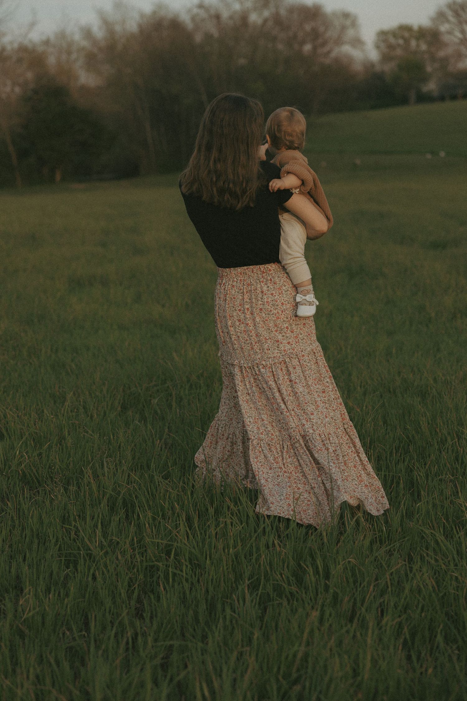 Mother in floral skirt carries baby through golden green field at sunset.