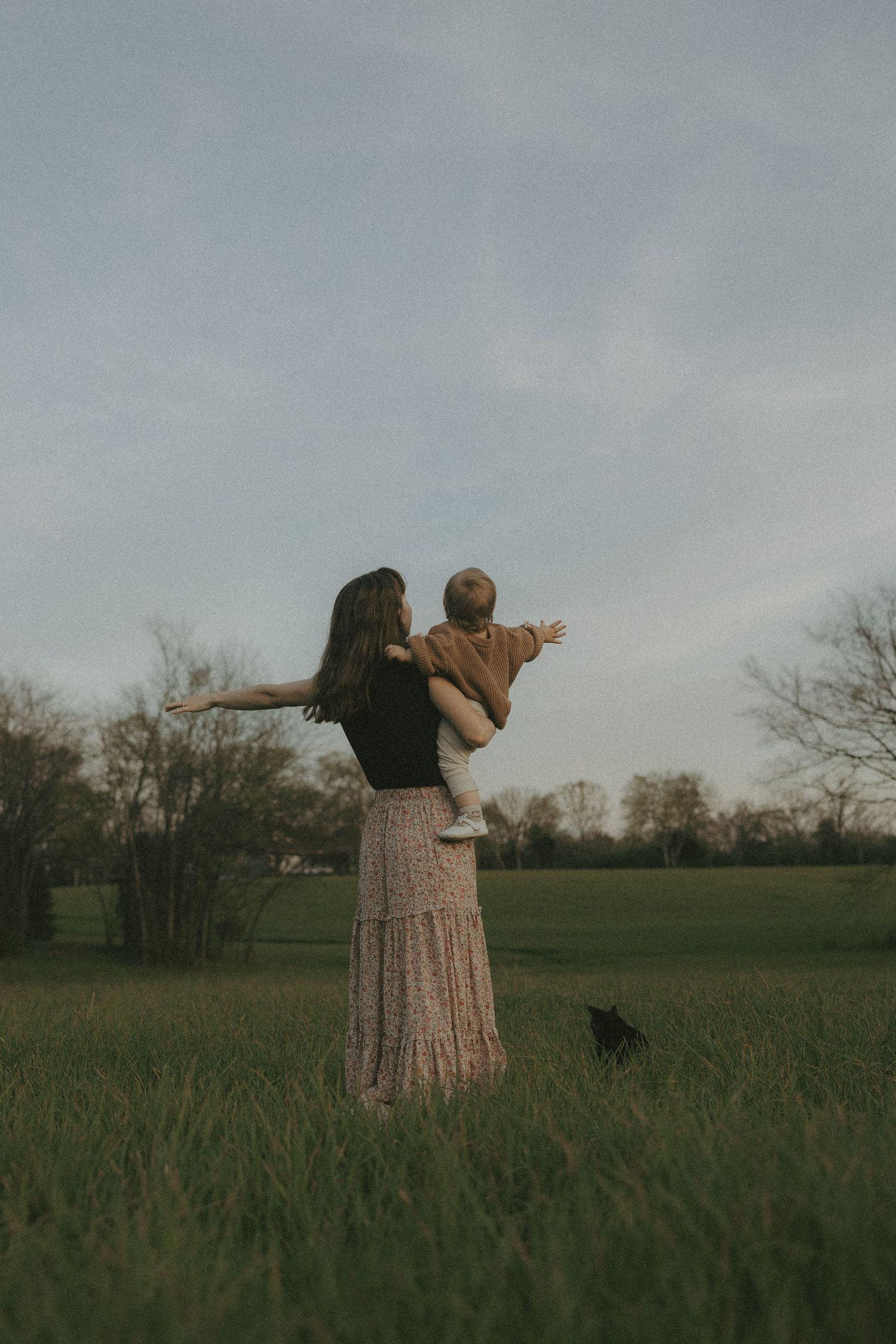 Mother lifts baby with outstretched arms in open green field at twilight.