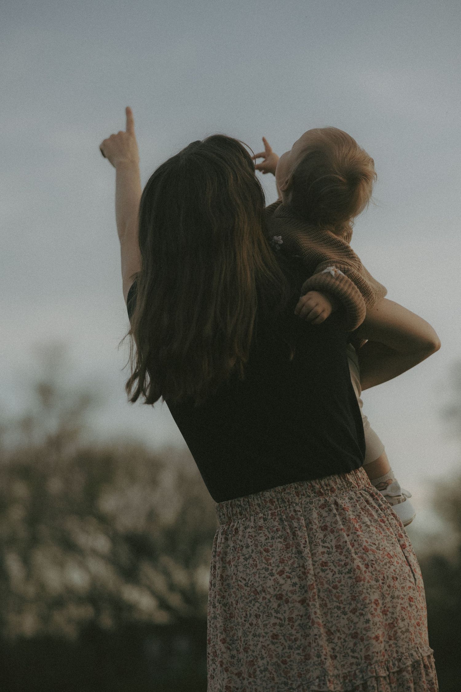 Mother points skyward while holding baby on hip in dusky outdoor setting.