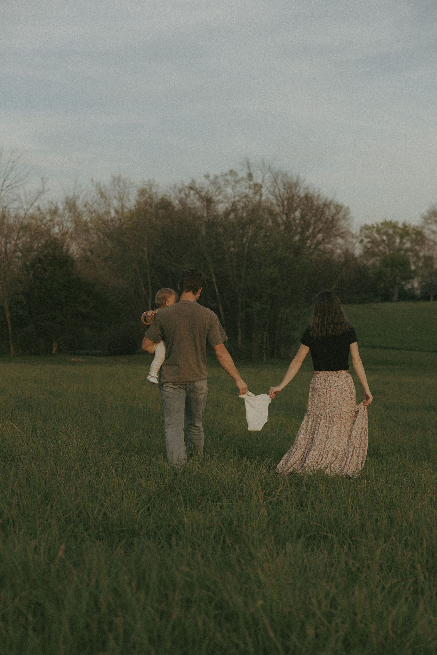 Family of three walks hand-in-hand through green meadow at dusk with trees behind.