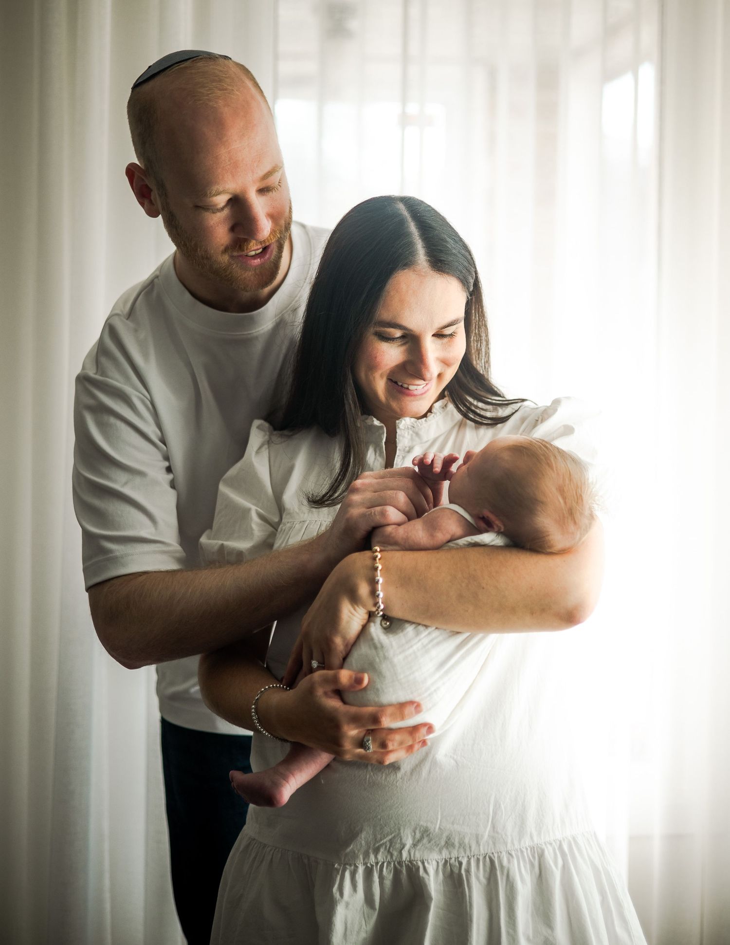 A tender family moment as parents share a joyful interaction with their newborn baby near a sunlit window.