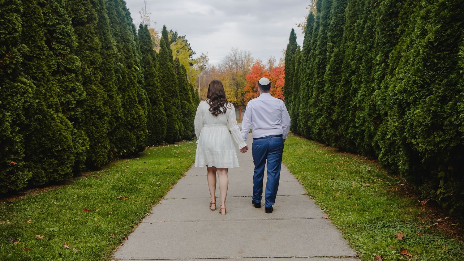 People stroll along a peaceful garden path lined with tall evergreen hedges on a cloudy day.