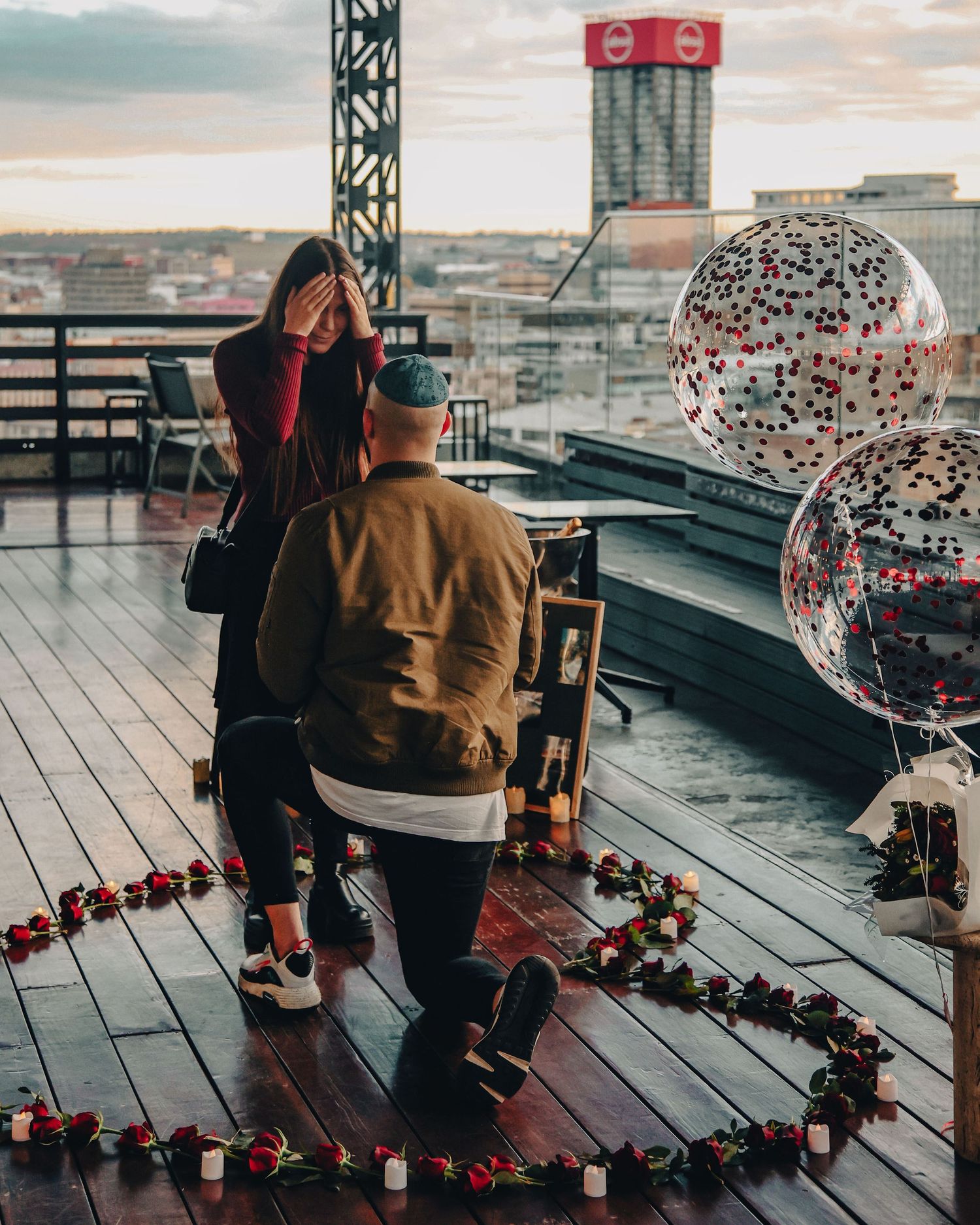 Romantic rooftop proposal setup with heart-shaped lights and disco balls against a city skyline at sunset.