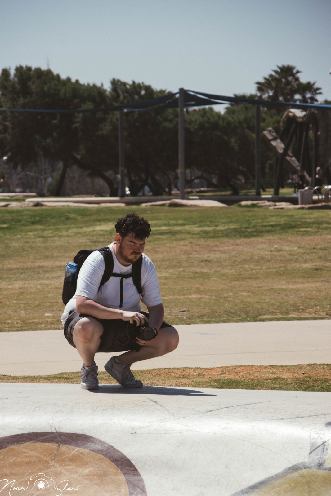 Person in casual attire crouching on a concrete surface in a park setting with trees in the background.