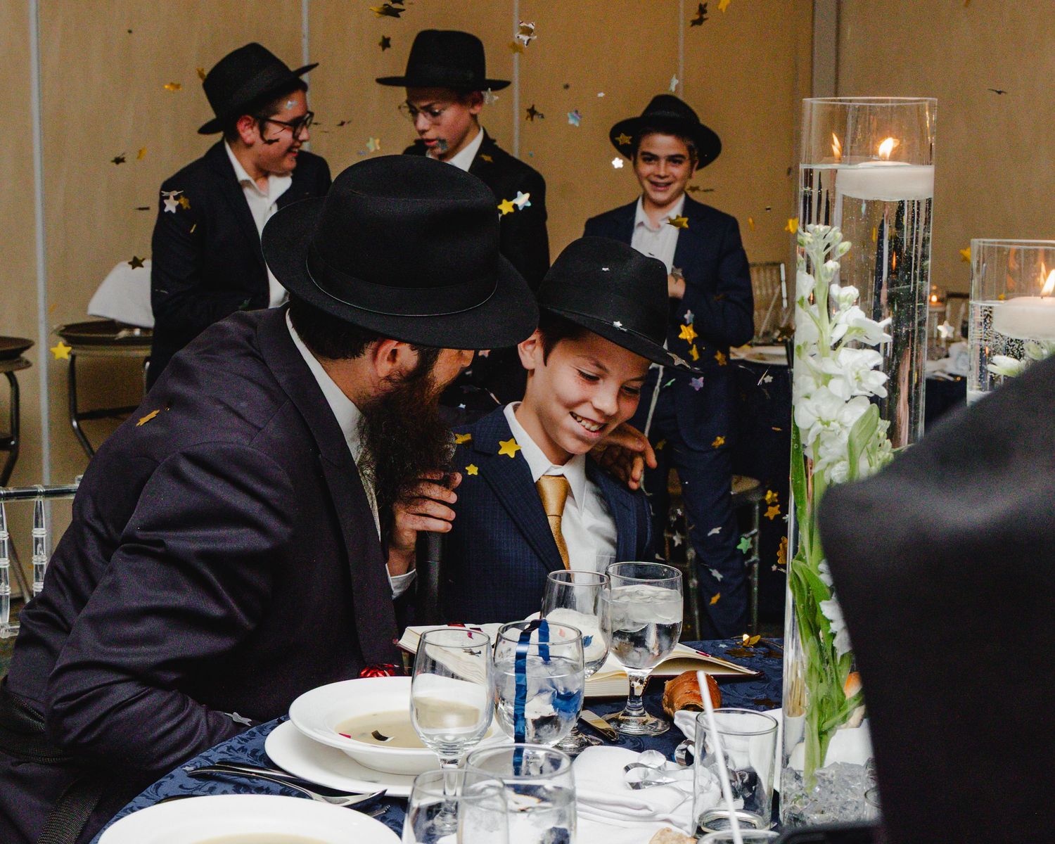 A group in formal attire with black hats sits at an elegant table with white linens and floral centerpieces.
