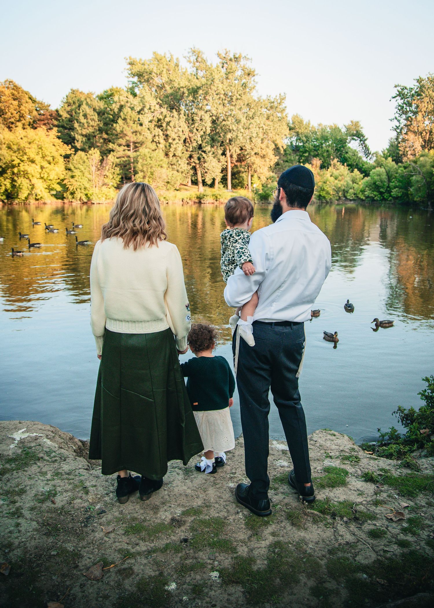 Family silhouettes standing by a lake at sunset watching ducks swim in golden evening light.