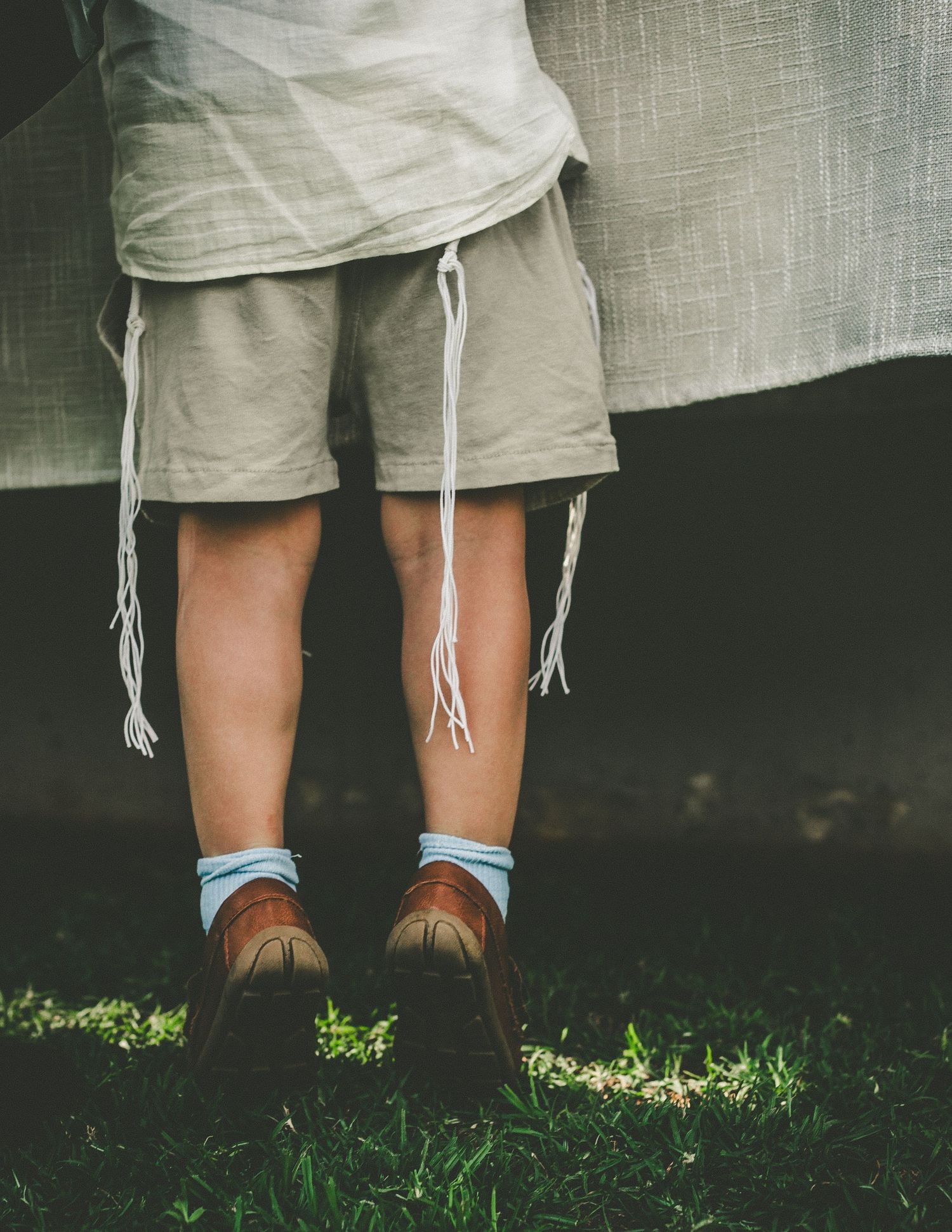 Close-up view of legs wearing khaki shorts with drawstrings, light blue socks, and brown leather shoes on grass.