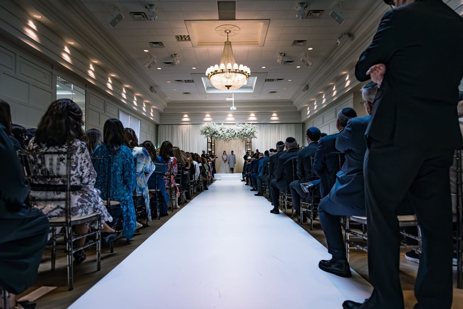 A grand ballroom with crystal chandeliers and white carpet aisle is set up for an elegant indoor wedding ceremony.