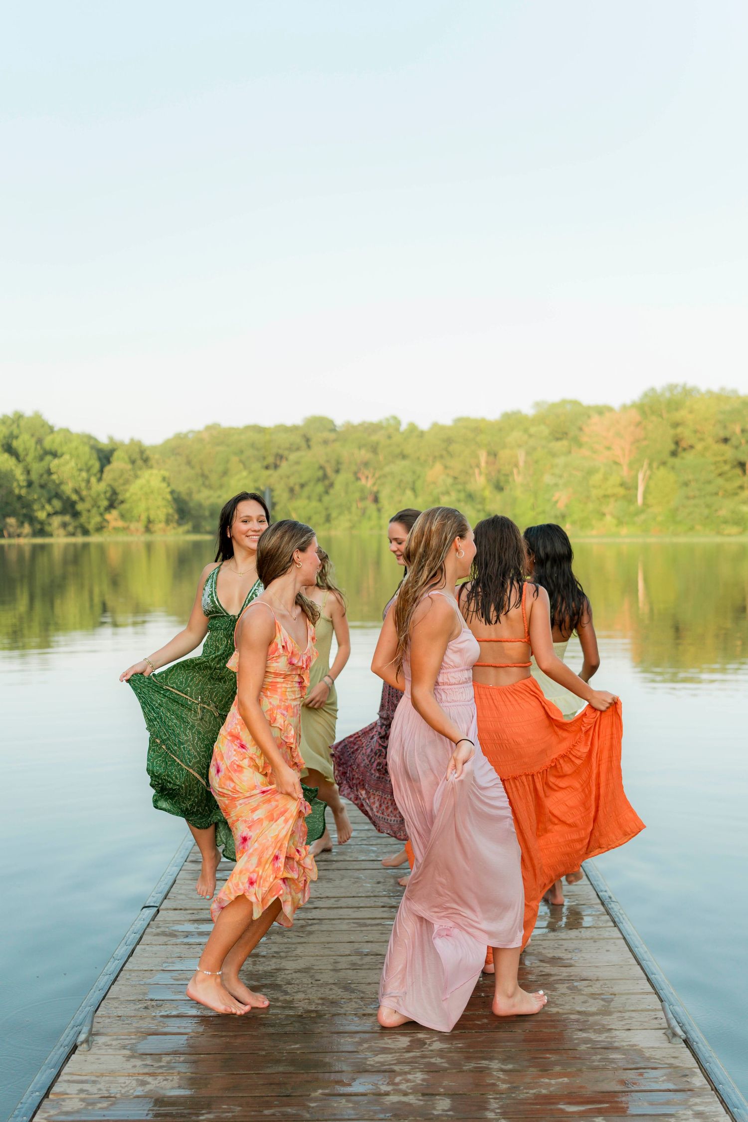A group in colorful dresses stands on a wooden dock by a lake, posing playfully in a sequence of three photos.