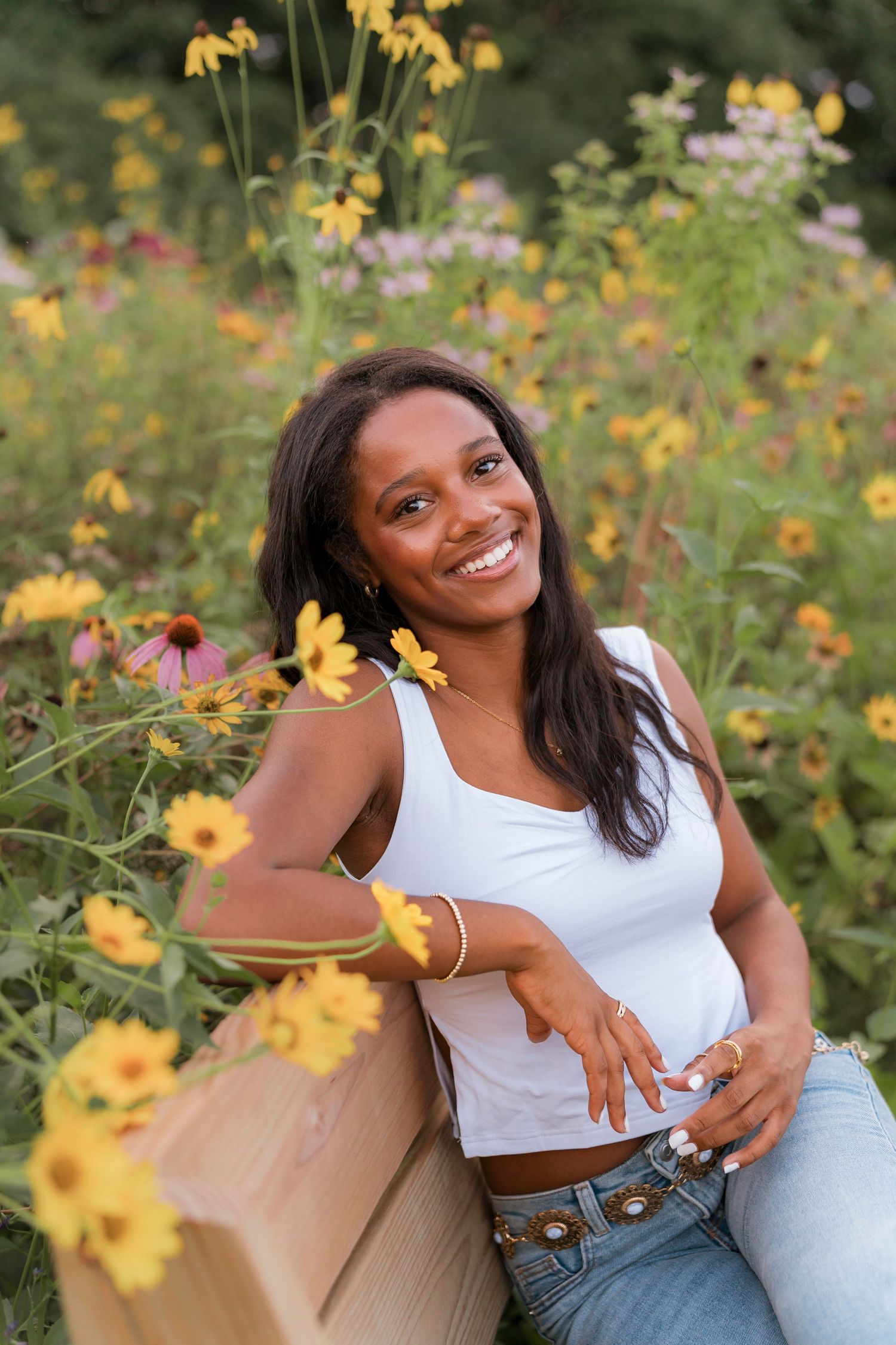 A person poses joyfully in a garden filled with vibrant yellow flowers and greenery on a sunny day.