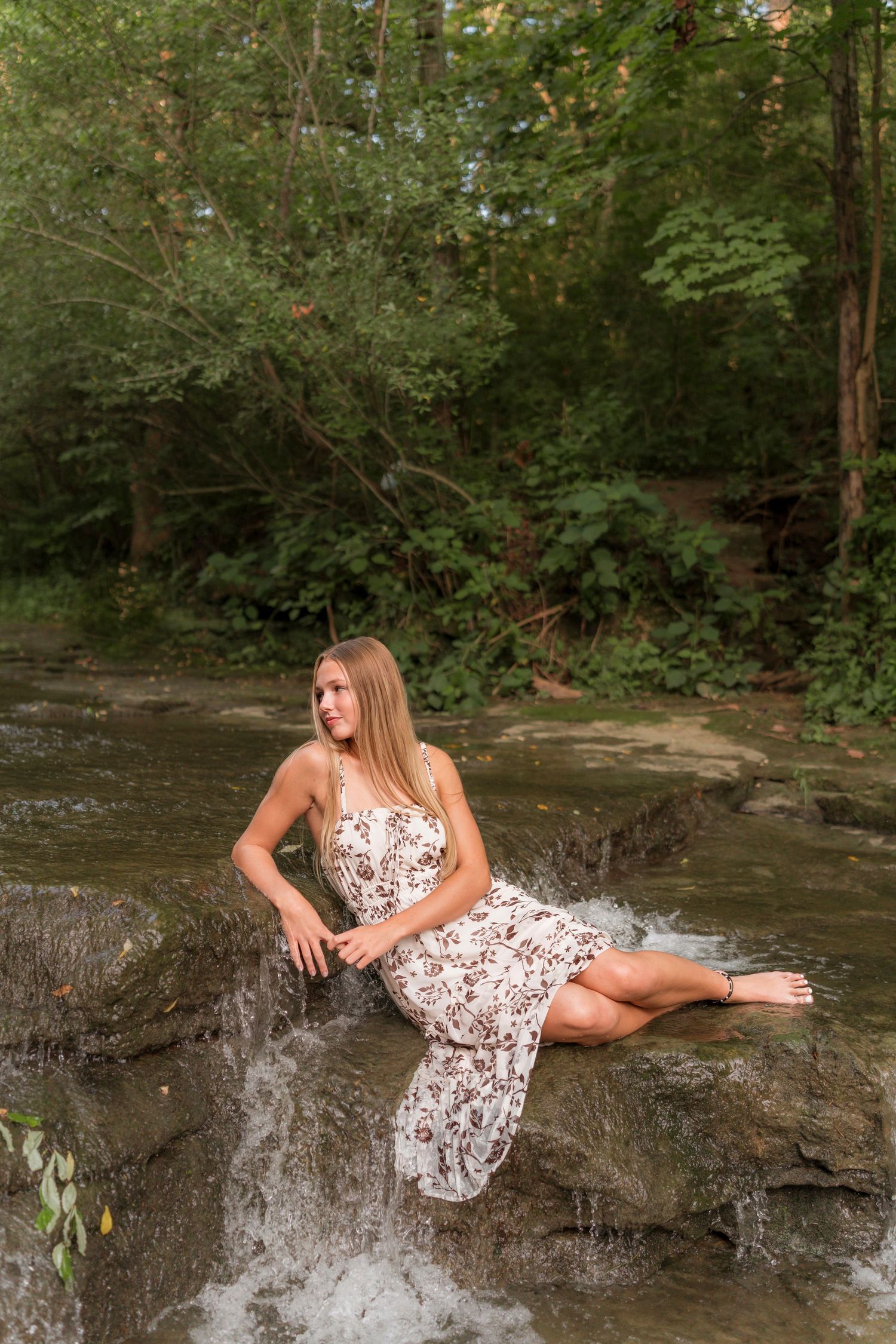 A person in a floral dress sits on rocks by a waterfall in a lush forest setting.