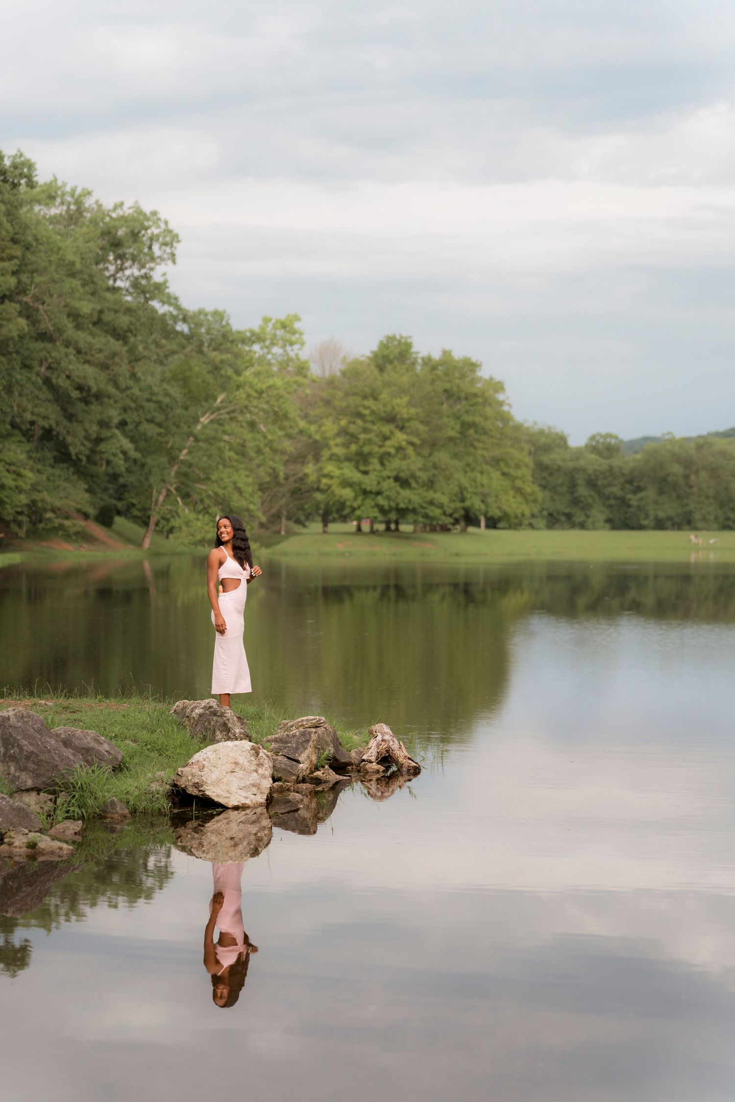 A person in a white dress stands on rocks by a serene lake, creating a perfect reflection in the still water.