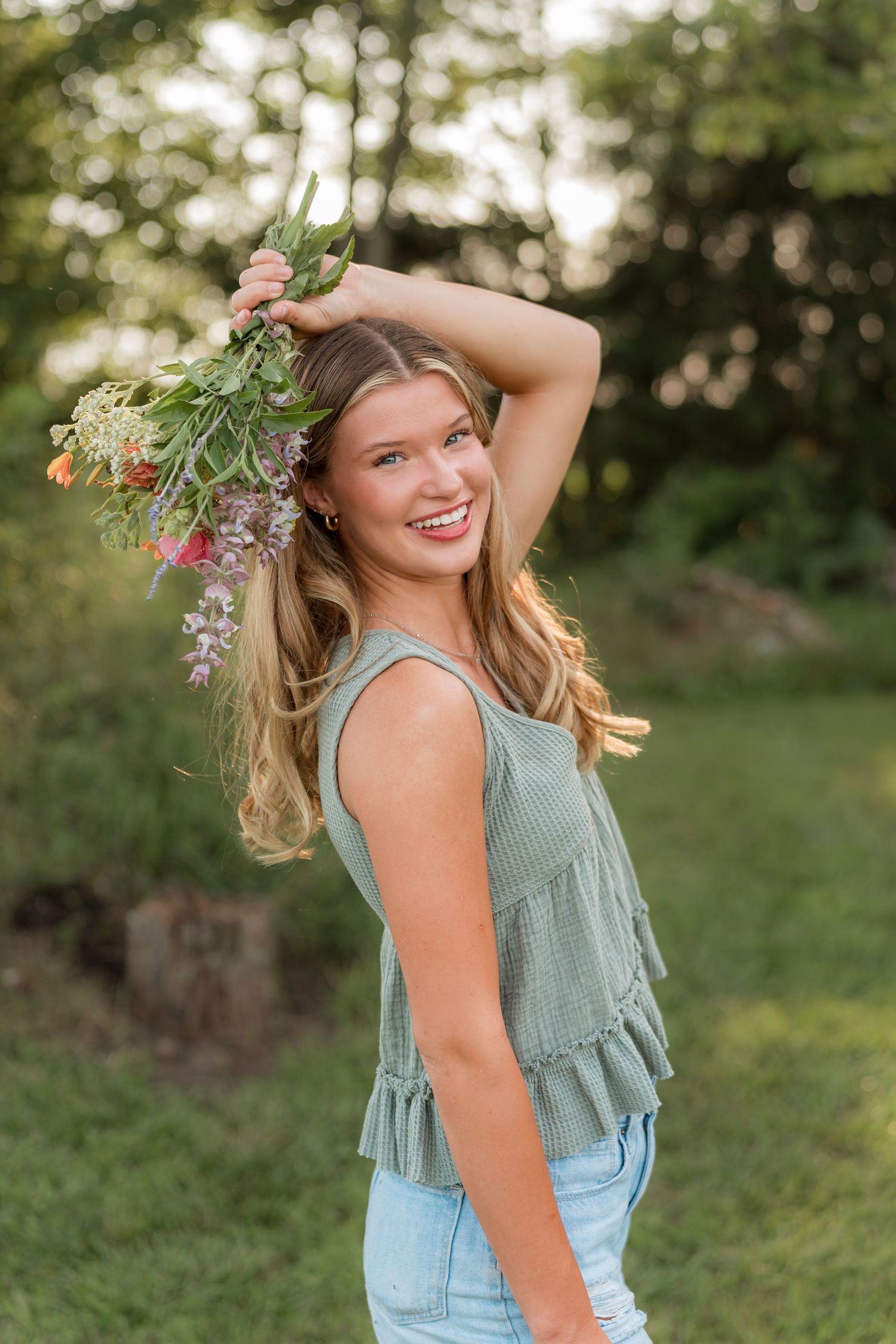 A woman in a mint green sleeveless top and jeans poses playfully outdoors in a garden setting.