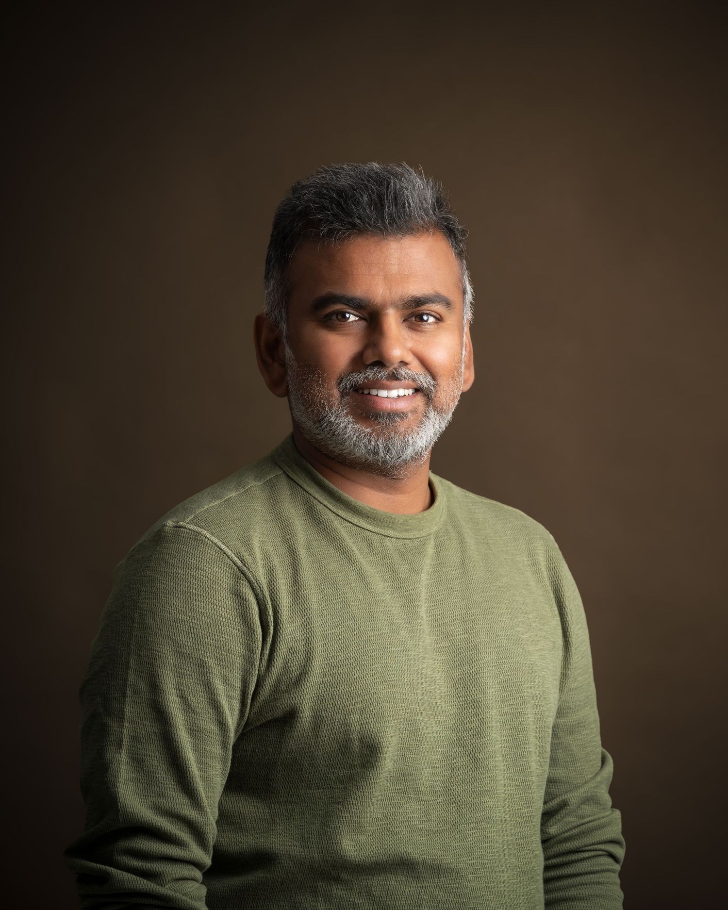 Professional studio portrait of someone wearing an olive green sweater against a dark brown background with dramatic lighting.