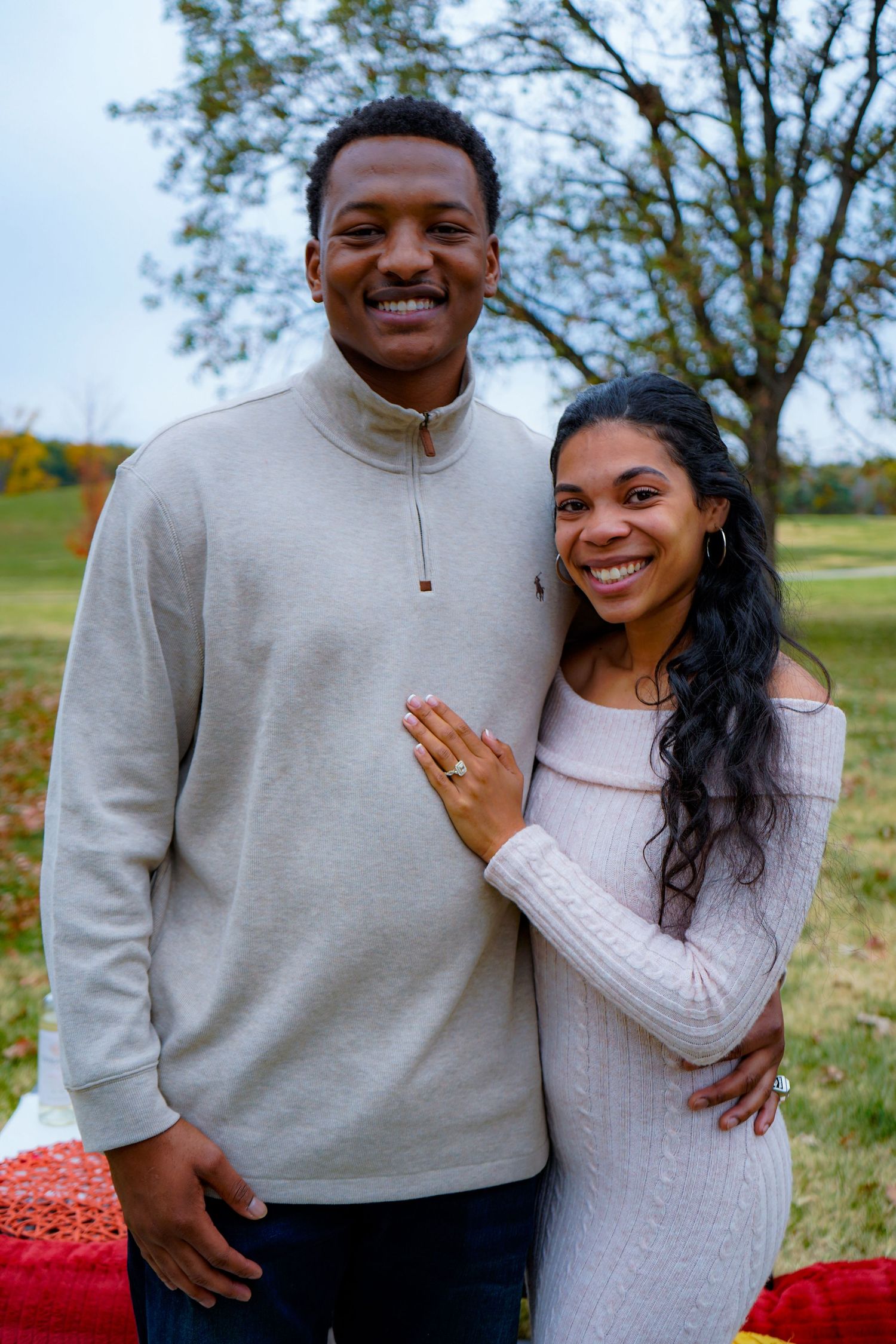 A couple in coordinating light-colored sweaters pose together outdoors during an autumn engagement photo session.