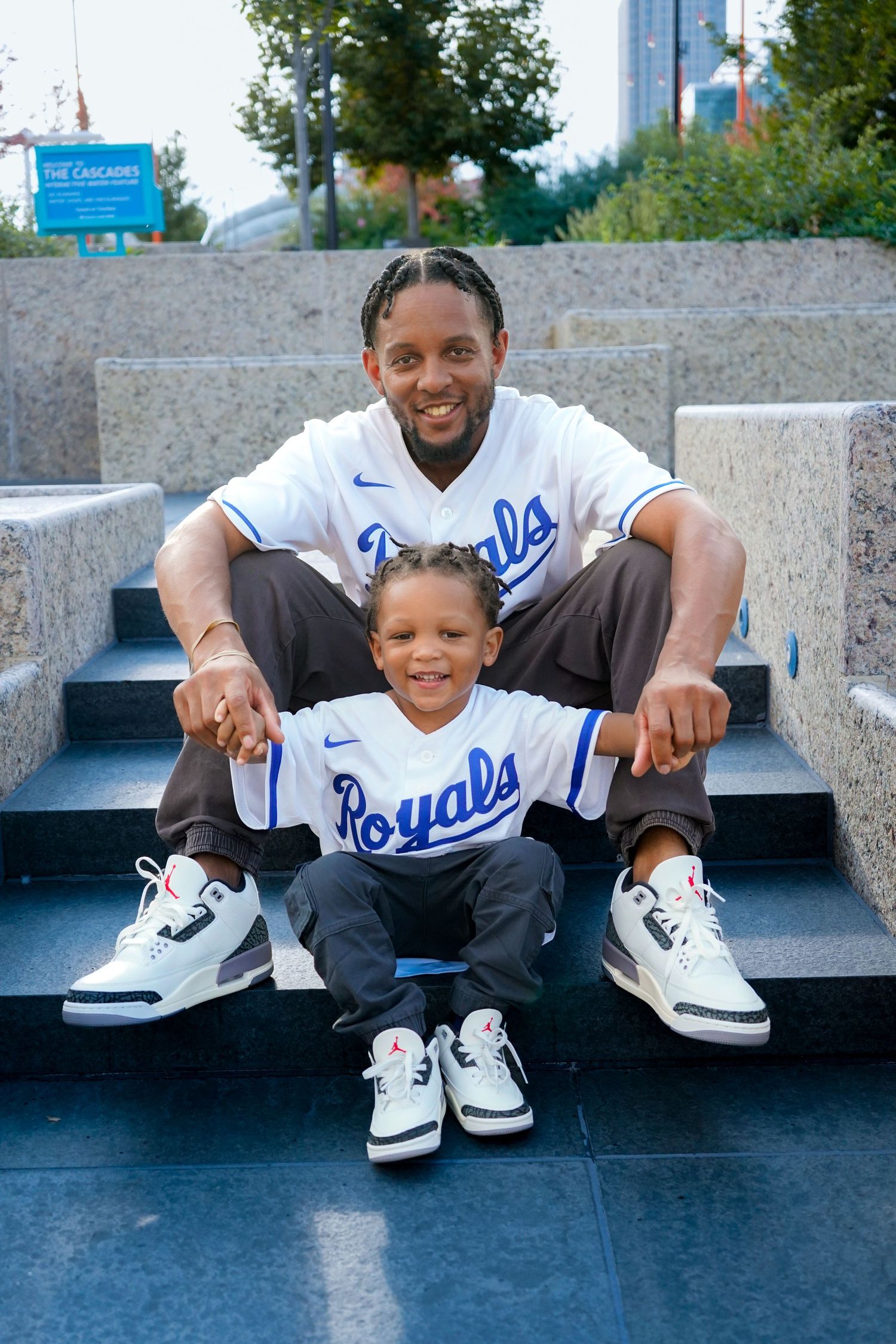 Two people wearing Kansas City Royals jerseys and matching white sneakers sit together on concrete steps outdoors.