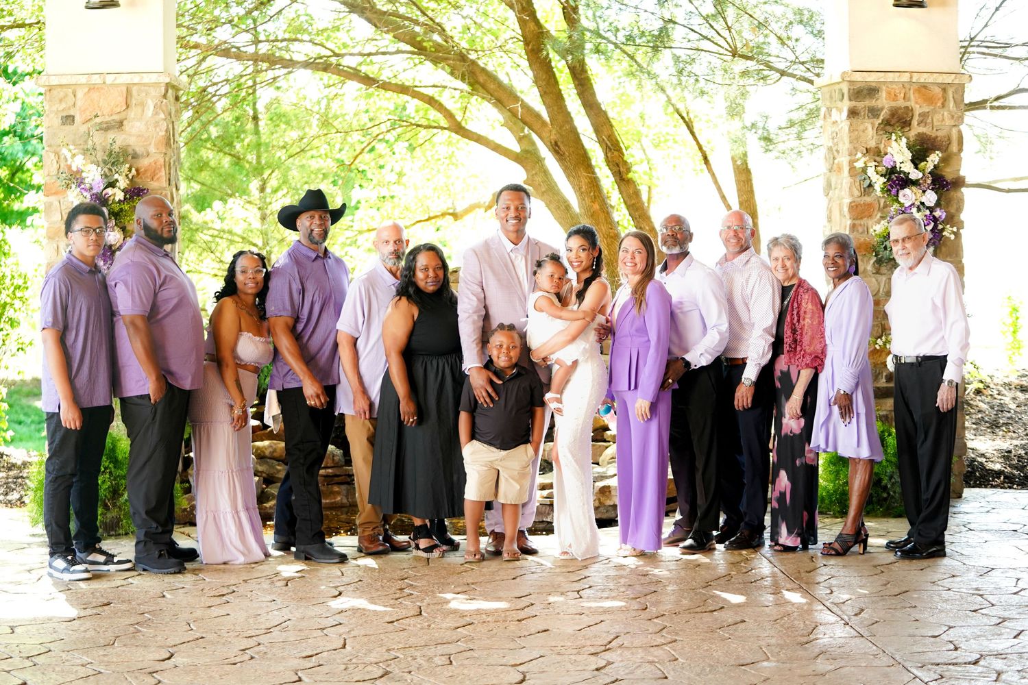 Large wedding party gathers under trees for a panoramic group photo.