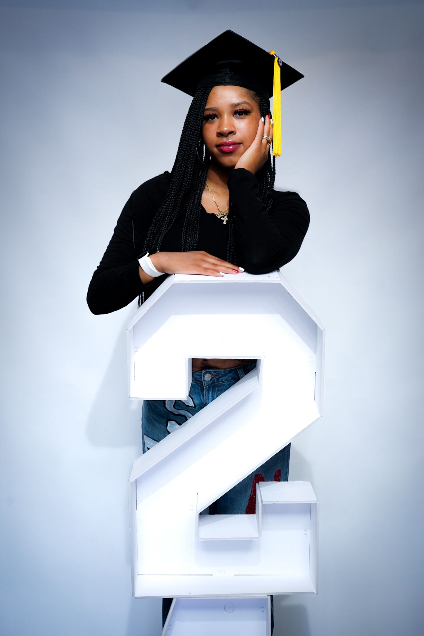 Graduate in black attire and cap poses with large white number 2 against light background.
