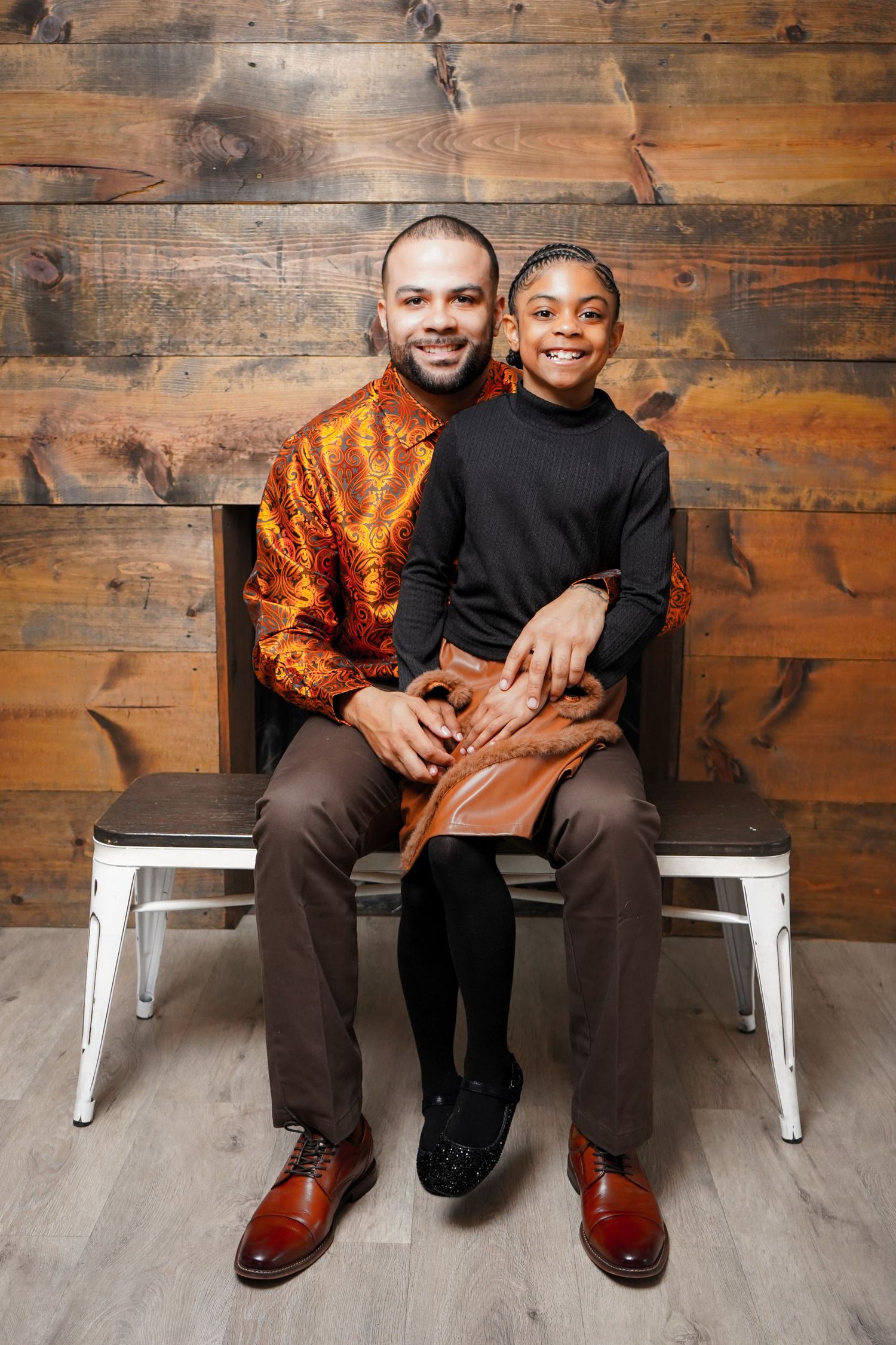 Two people sit close together on white chairs against a rustic wooden wall background.